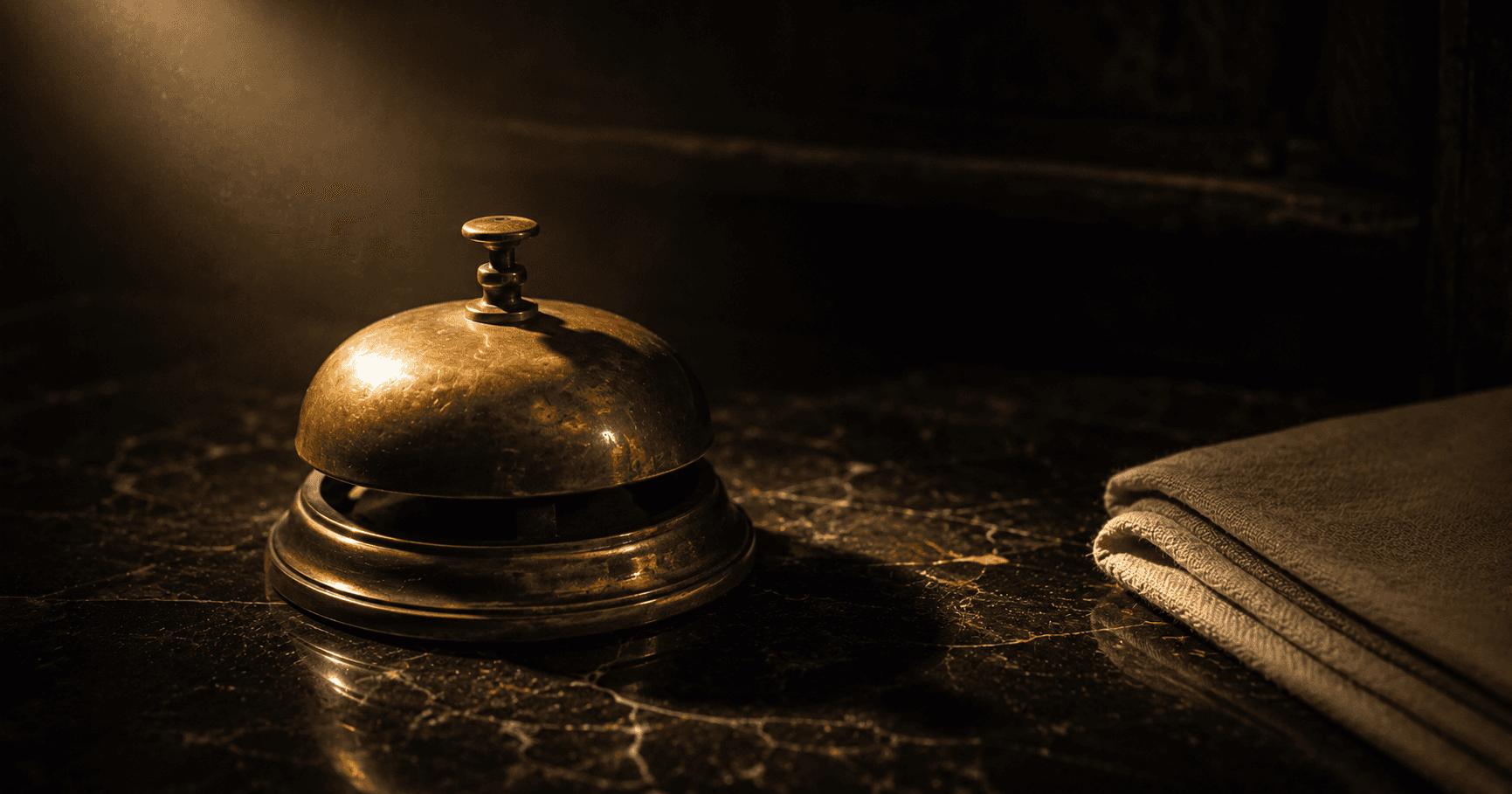 A vintage brass desk bell on dark polished marble, lit by a single warm directional beam — attentive service waiting on the patient's terms.